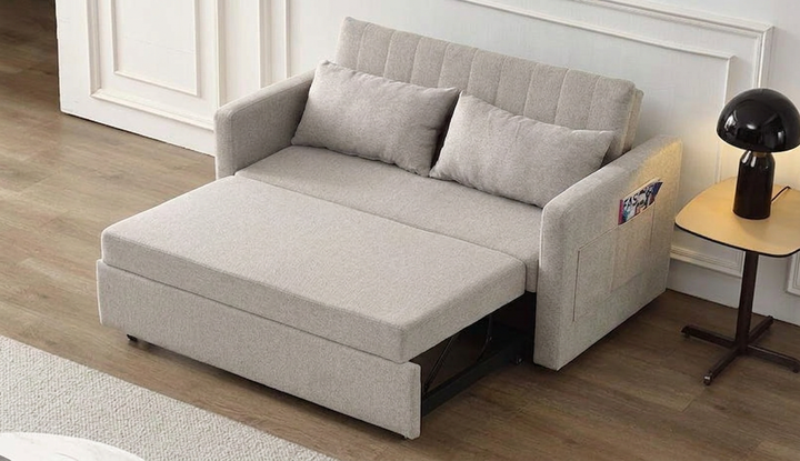 Sofá cama beige en el salón, desplegado en forma de cama con dos cojines. Decoración moderna con suelo de madera, mesita auxiliar y lámpara negra. Ambiente acogedor.