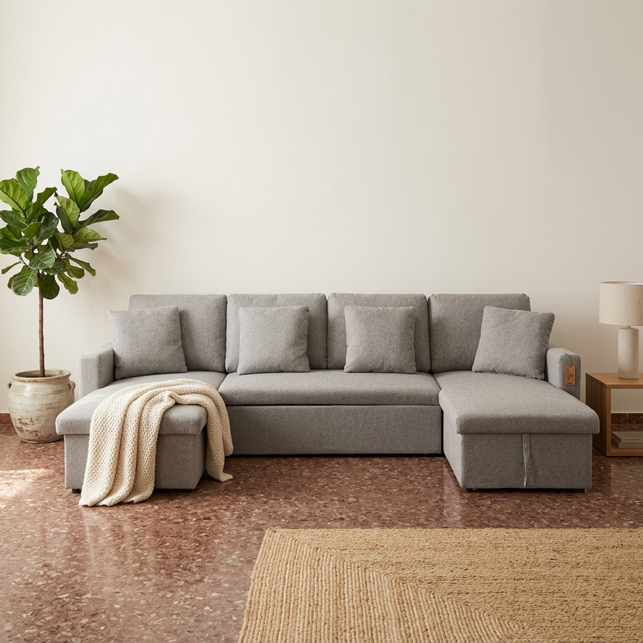 Un salón acogedor y moderno cuenta con un sofá modular gris cubierto con una manta de punto beige. Una planta en maceta y una lámpara aportan calidez al suelo de terracota.