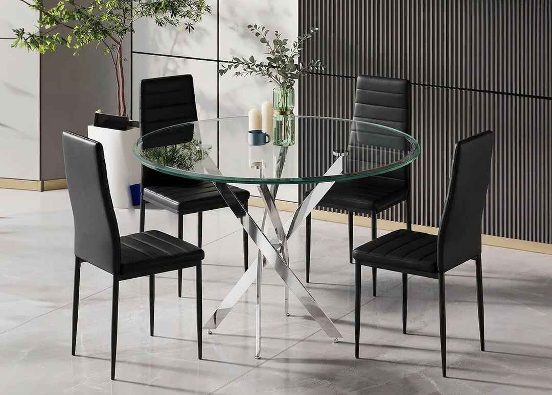Comedor moderno con una mesa redonda de cristal sobre una elegante base metálica, rodeada de cuatro sillas negras. Un jarrón con ramas verdes y velas adorna la mesa.