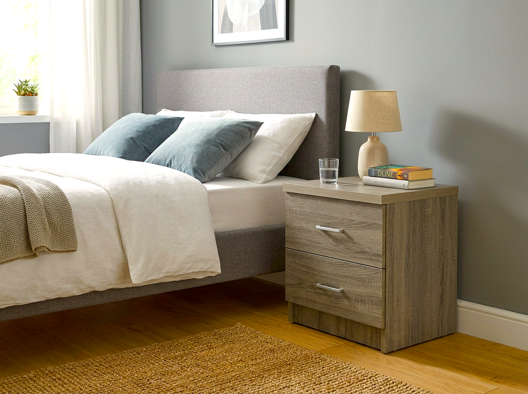 Un dormitorio acogedor con una cama tapizada en gris, ropa de cama blanca y cojines azul grisáceos. En una mesita de noche de madera hay una lámpara, un libro y un vaso.