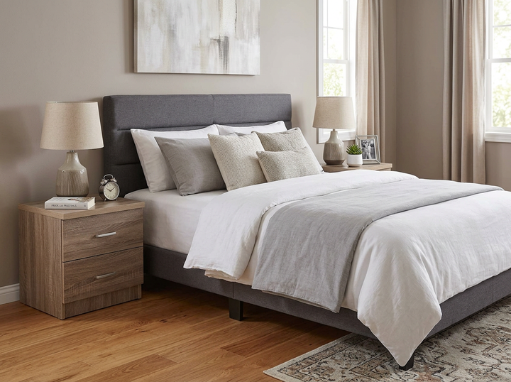 Dormitorio moderno con una cama tapizada en gris, ropa de cama blanca y gris, y mesitas de noche con lámparas. La luz del sol se filtra a través de las cortinas beige. Ambiente acogedor.