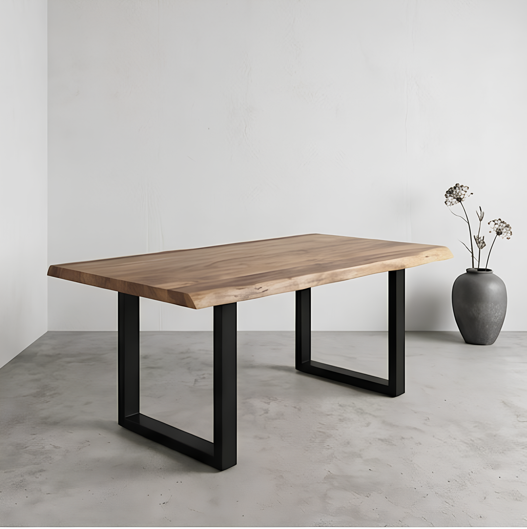 En una habitación minimalista, con el suelo y las paredes grises, hay una mesa de madera con patas de metal negro; en una esquina hay un jarrón gris con flores secas.