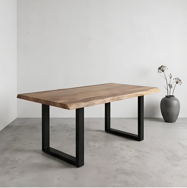 En una habitación minimalista, con el suelo y las paredes grises, hay una mesa de madera con patas de metal negro; en una esquina hay un jarrón gris con flores secas.