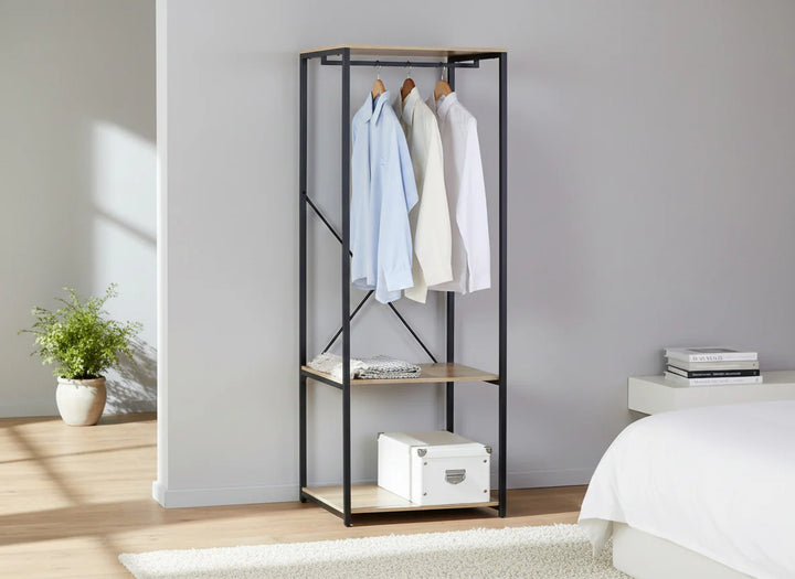 Escena minimalista de un dormitorio con un elegante armario abierto en el que cuelgan tres camisas perfectamente ordenadas. En la habitación, luminosa y espaciosa, hay una planta en maceta y algunos libros.