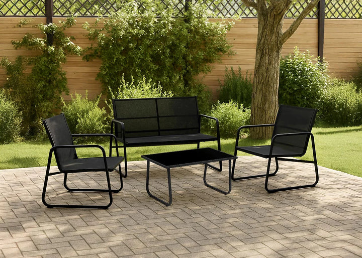 Un moderno conjunto de jardín compuesto por un banco negro, dos sillas y una mesa rectangular sobre una superficie de ladrillo, rodeado de vegetación y un árbol.