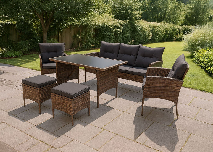 Conjunto de muebles de exterior con una mesa con tablero de cristal, sillas acolchadas y pufs en un patio de piedra. Los arbustos verdes y la luz del sol crean un ambiente sereno.