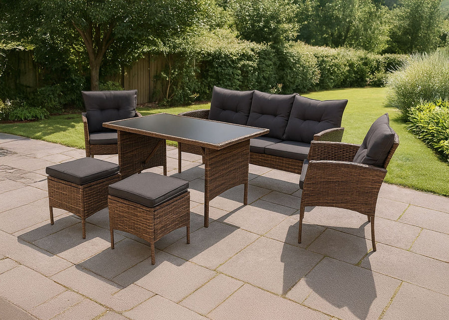 Conjunto de muebles de exterior con una mesa con tablero de cristal, sillas acolchadas y pufs en un patio de piedra. Los arbustos verdes y la luz del sol crean un ambiente sereno.