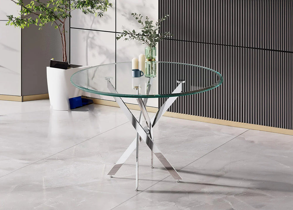 Comedor moderno con una mesa redonda de cristal sobre patas de metal brillante. Cerca, sobre el suelo de baldosas, hay una planta en maceta y algunos objetos decorativos. Minimalista y elegante.