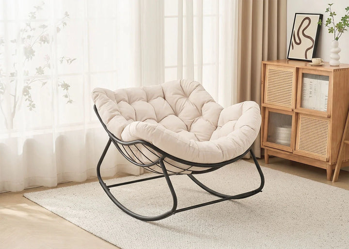 Una acogedora mecedora de color beige con cojín capitoné, situada sobre una alfombra clara. Se encuentra junto a un armario de madera con libros y una planta, bañada por la suave luz del sol.
