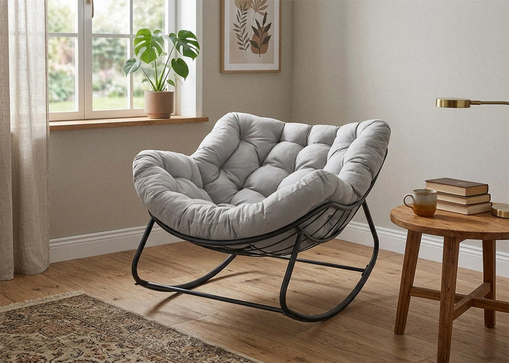 Una acogedora habitación con un lujoso sillón gris capitoné sobre una estructura de metal negro. Una mesita auxiliar de madera con libros, té y una suave luz natural que entra por la ventana. Un ambiente relajante.