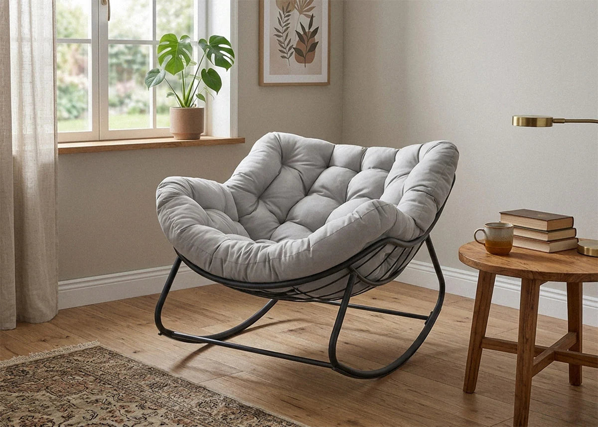 Una acogedora habitación con un lujoso sillón gris capitoné sobre una estructura de metal negro. Una mesita auxiliar de madera con libros, té y una suave luz natural que entra por la ventana. Un ambiente relajante.