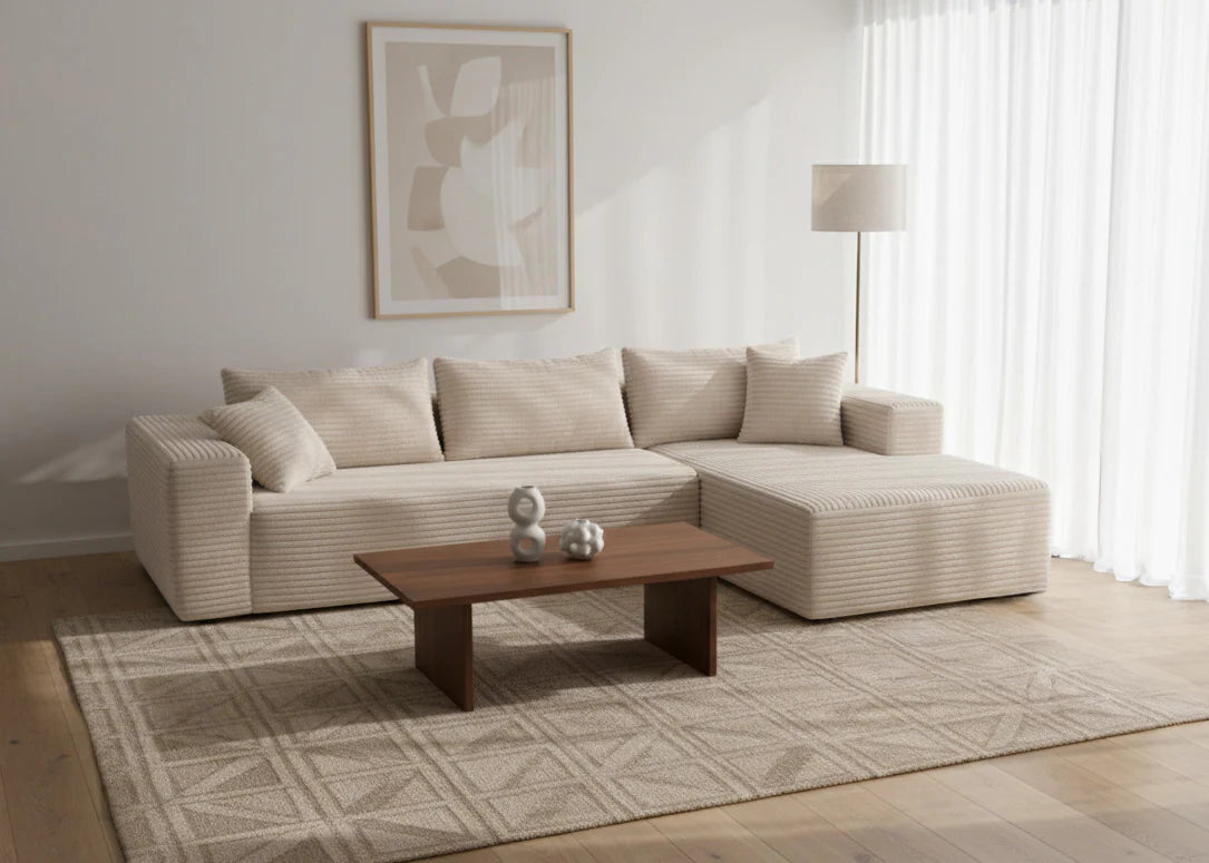 Salón minimalista con un sofá en forma de L de color crema, cuadros abstractos enmarcados, una mesa de centro de madera, una alfombra geométrica, una lámpara y cortinas transparentes. Ambiente tranquilo y elegante.