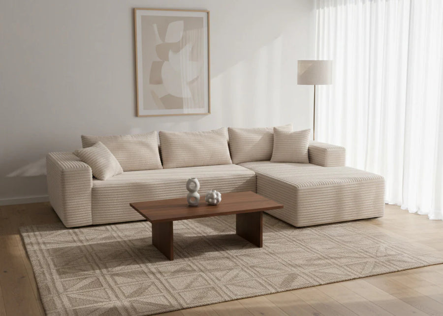 Salón minimalista con un sofá en forma de L de color crema, cuadros abstractos enmarcados, una mesa de centro de madera, una alfombra geométrica, una lámpara y cortinas transparentes. Ambiente tranquilo y elegante.