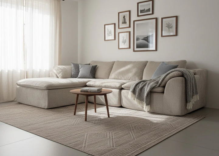 Un acogedor salón con un sofá modular de color beige, cojines azul grisáceos, una manta de punto, una mesa de centro de madera y cuadros enmarcados en una pared blanca. La suave luz del sol se filtra a través de las cortinas transparentes, creando un ambiente cálido y acogedor.