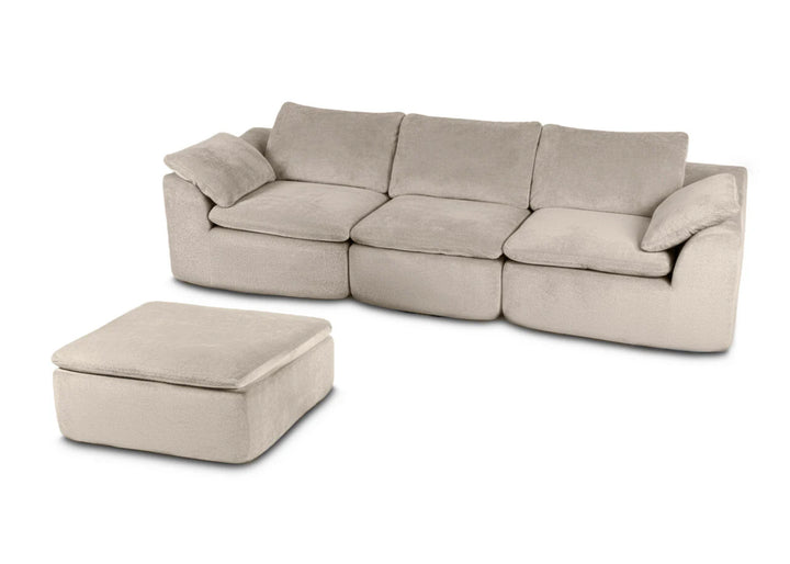 Un sofá modular de color gris claro y tejido mullido, con tres cojines y una otomana a juego, que transmite un ambiente acogedor y agradable.